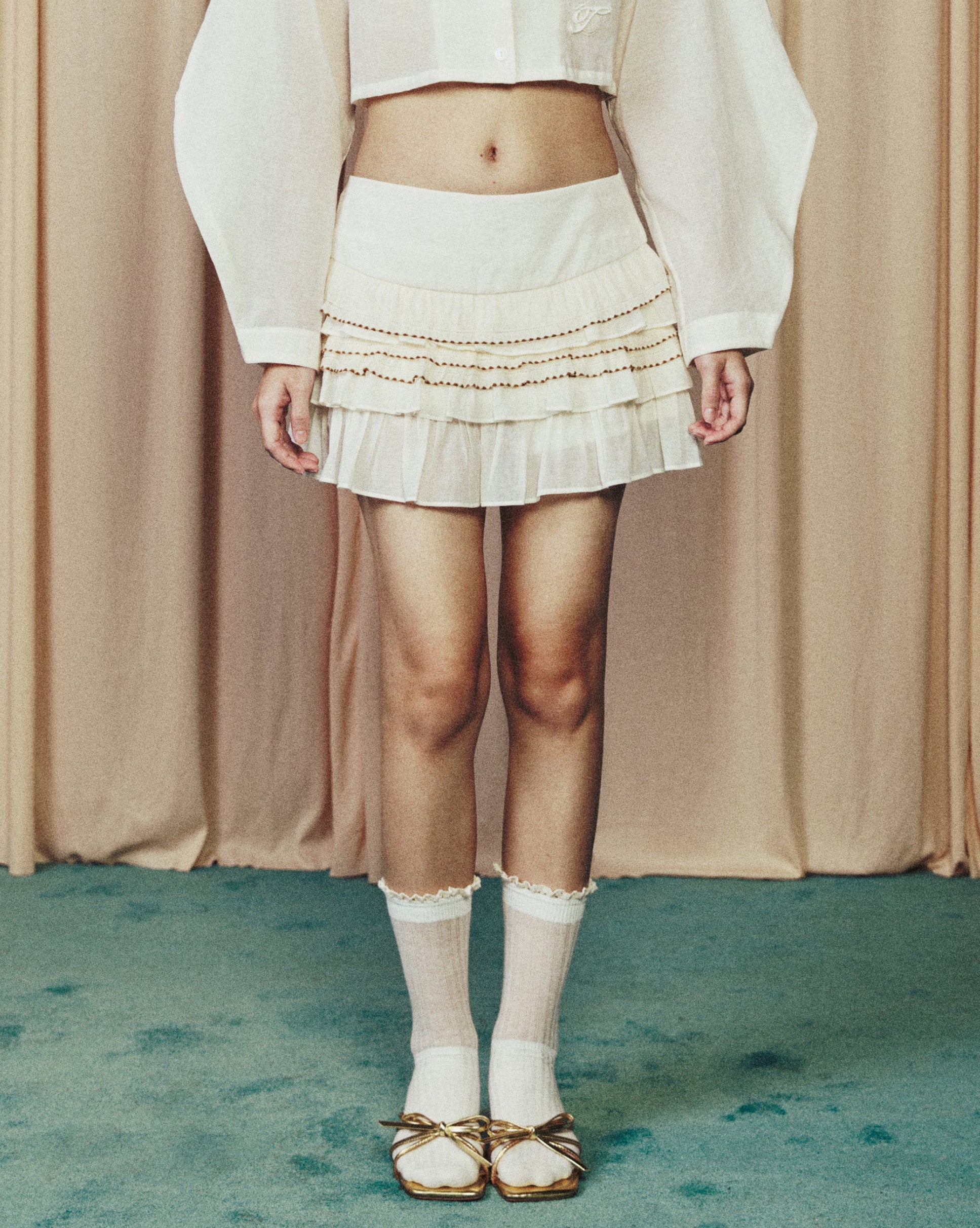 Person wearing a white outfit with a sheer top and skirt against a beige curtain background