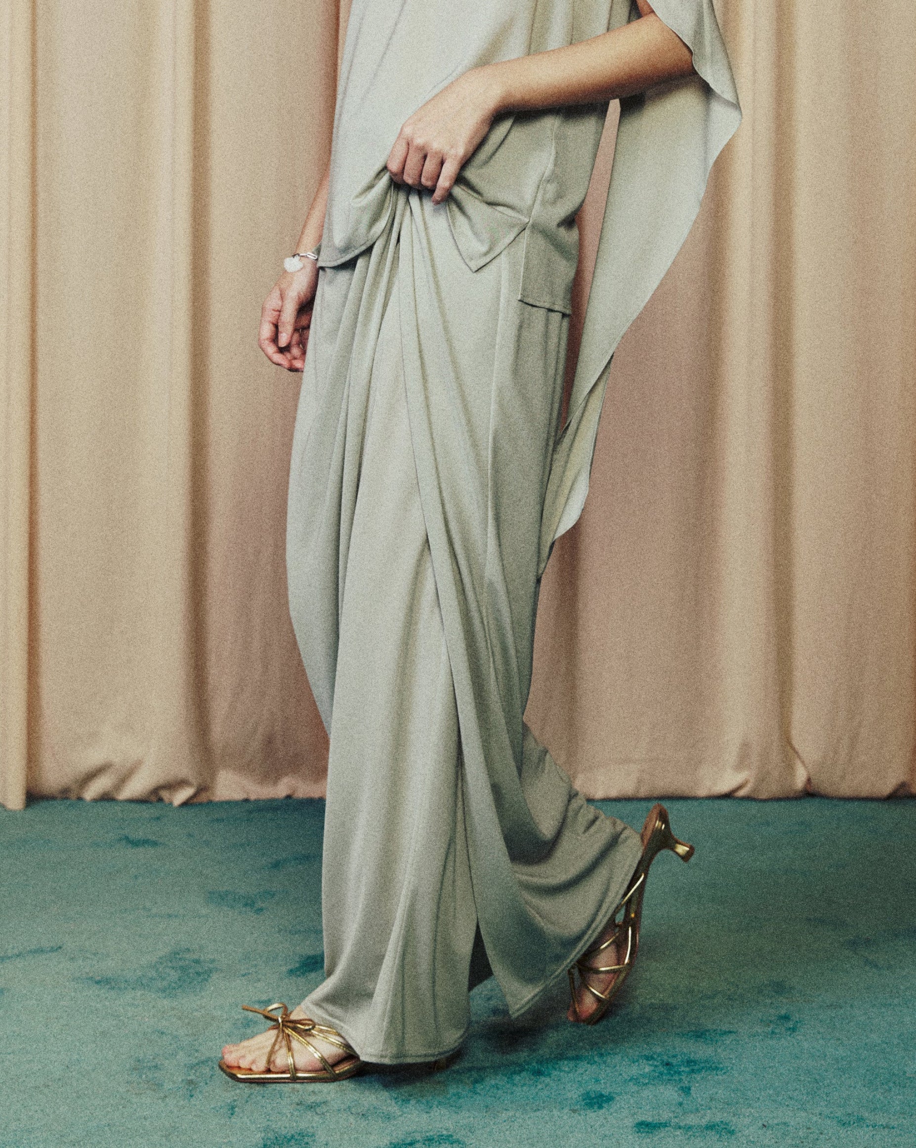Woman in a flowing light green dress standing in front of beige curtains.